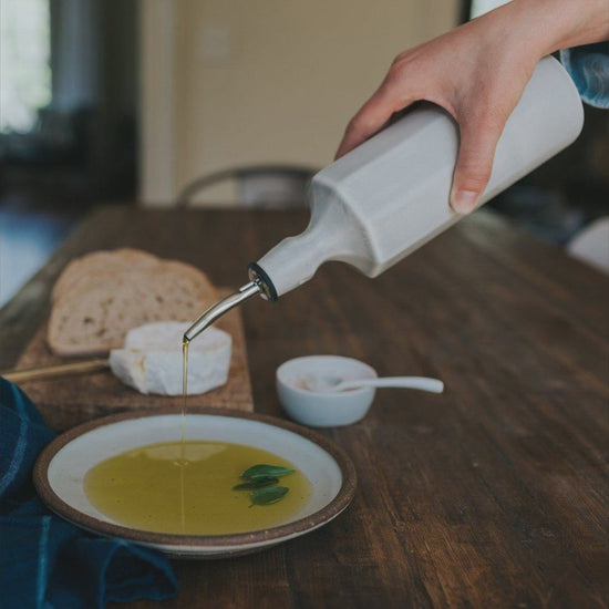 A hand pours olive oil from a porcelain dispenser into a bowl on a table