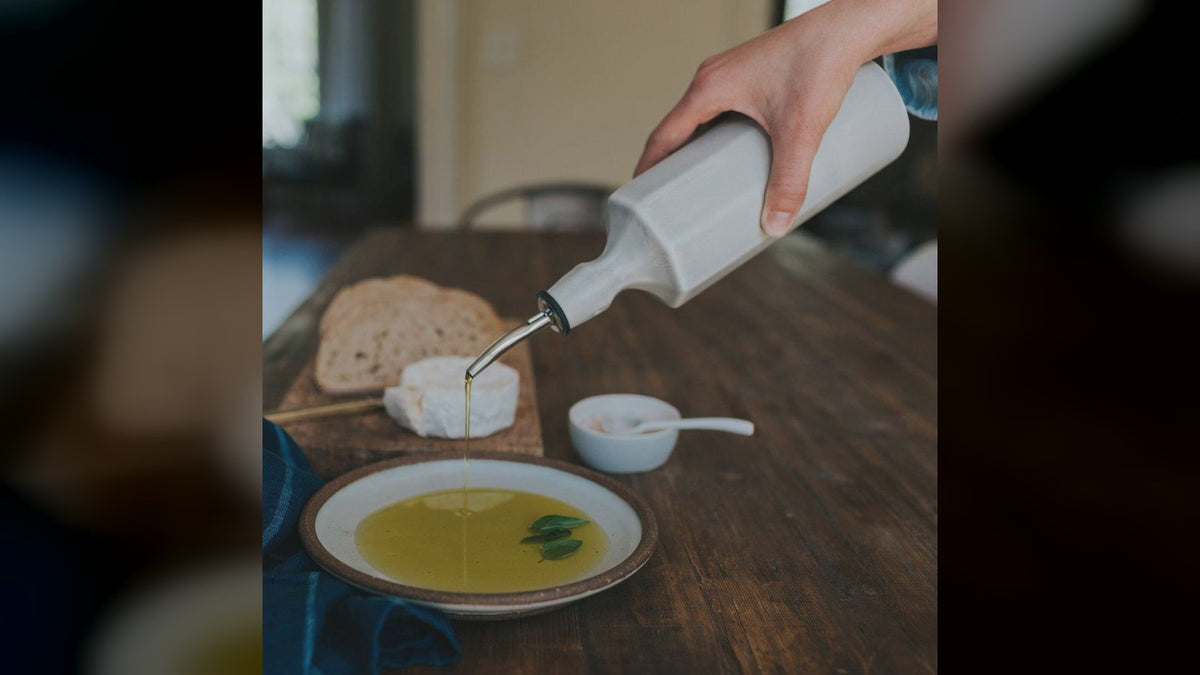 A hand pours olive oil from a porcelain dispenser into a bowl on a table