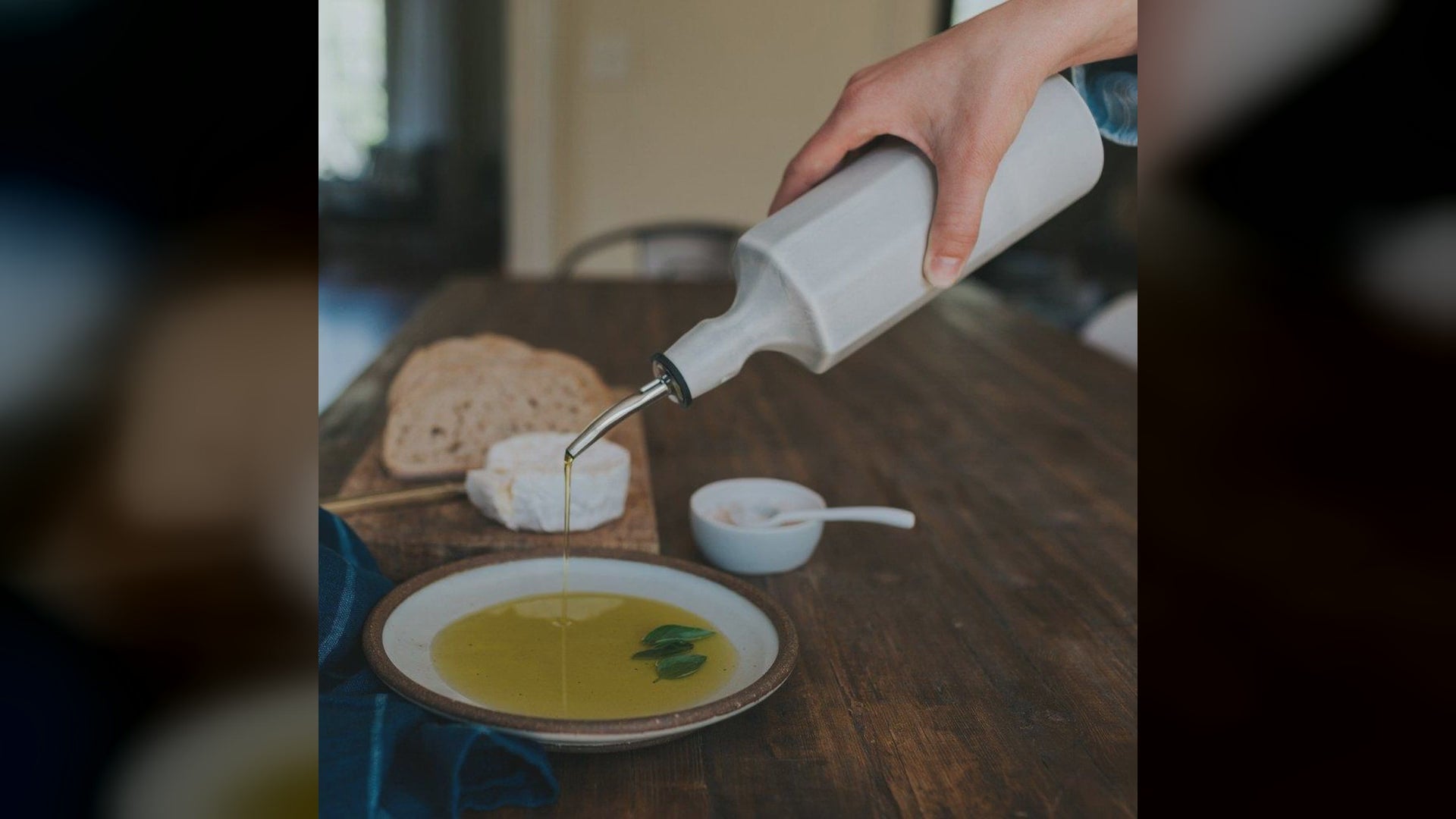 A hand pours olive oil from a porcelain dispenser into a bowl on a table